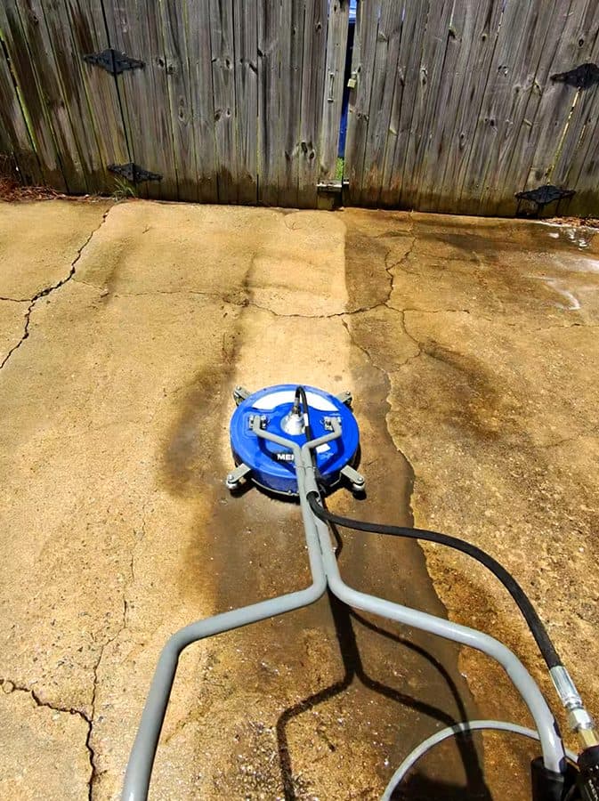 Pressure washing surface cleaner on stained concrete beside a wood fence